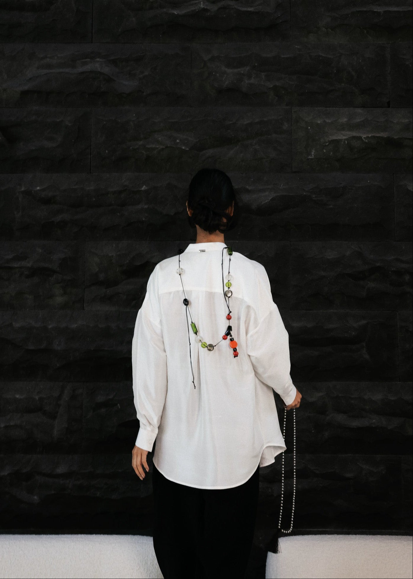 Person wearing a white shirt and dark pants against a black textured wall.