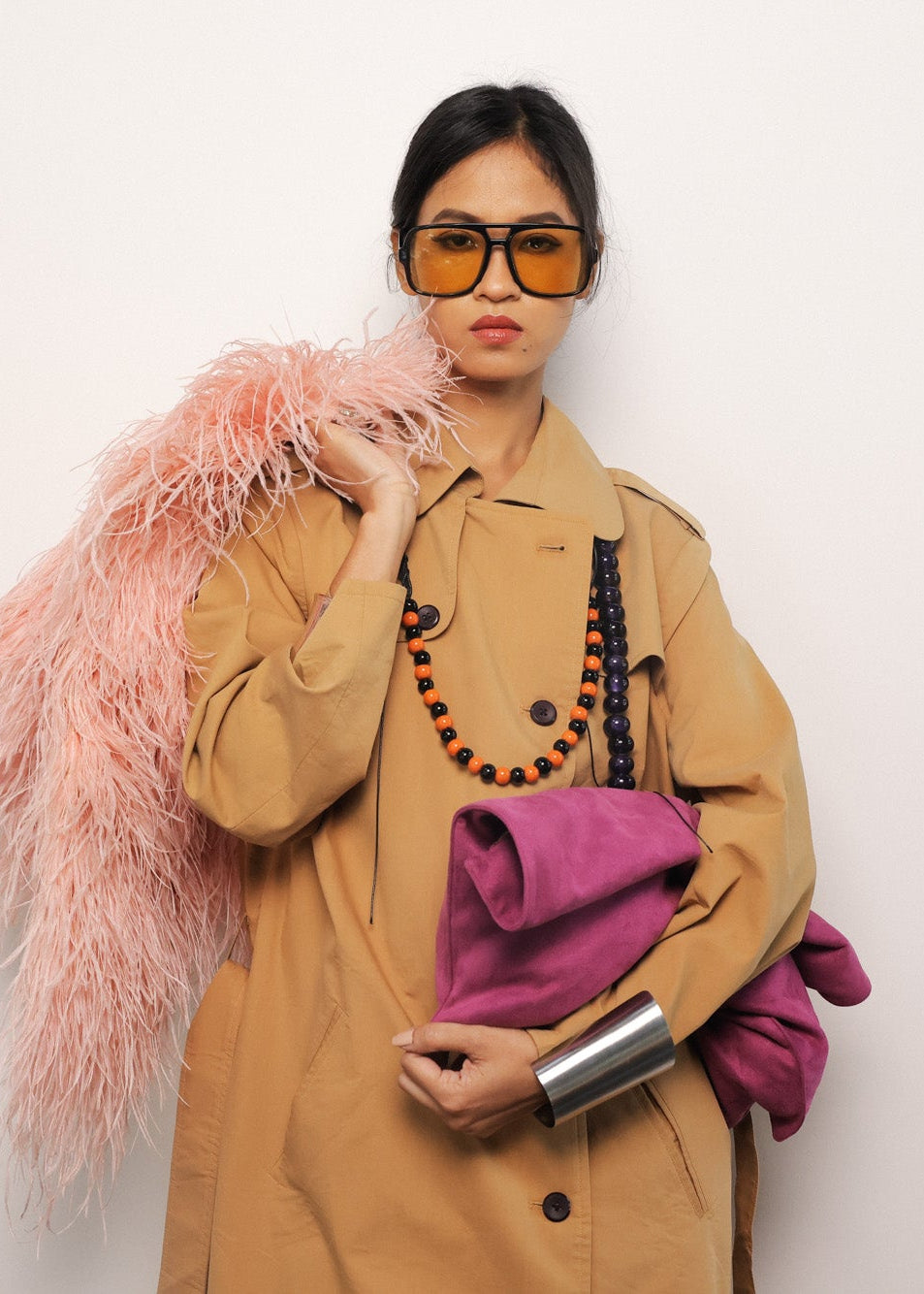 Person wearing a beige trench coat with a pink fur stole and holding a pink bag against a white background