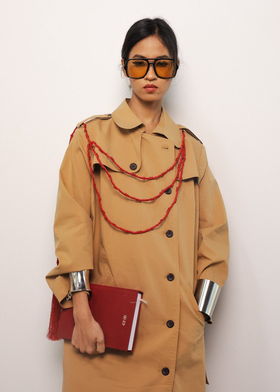 Person wearing a beige trench coat with a red book and decorative strings against a white background