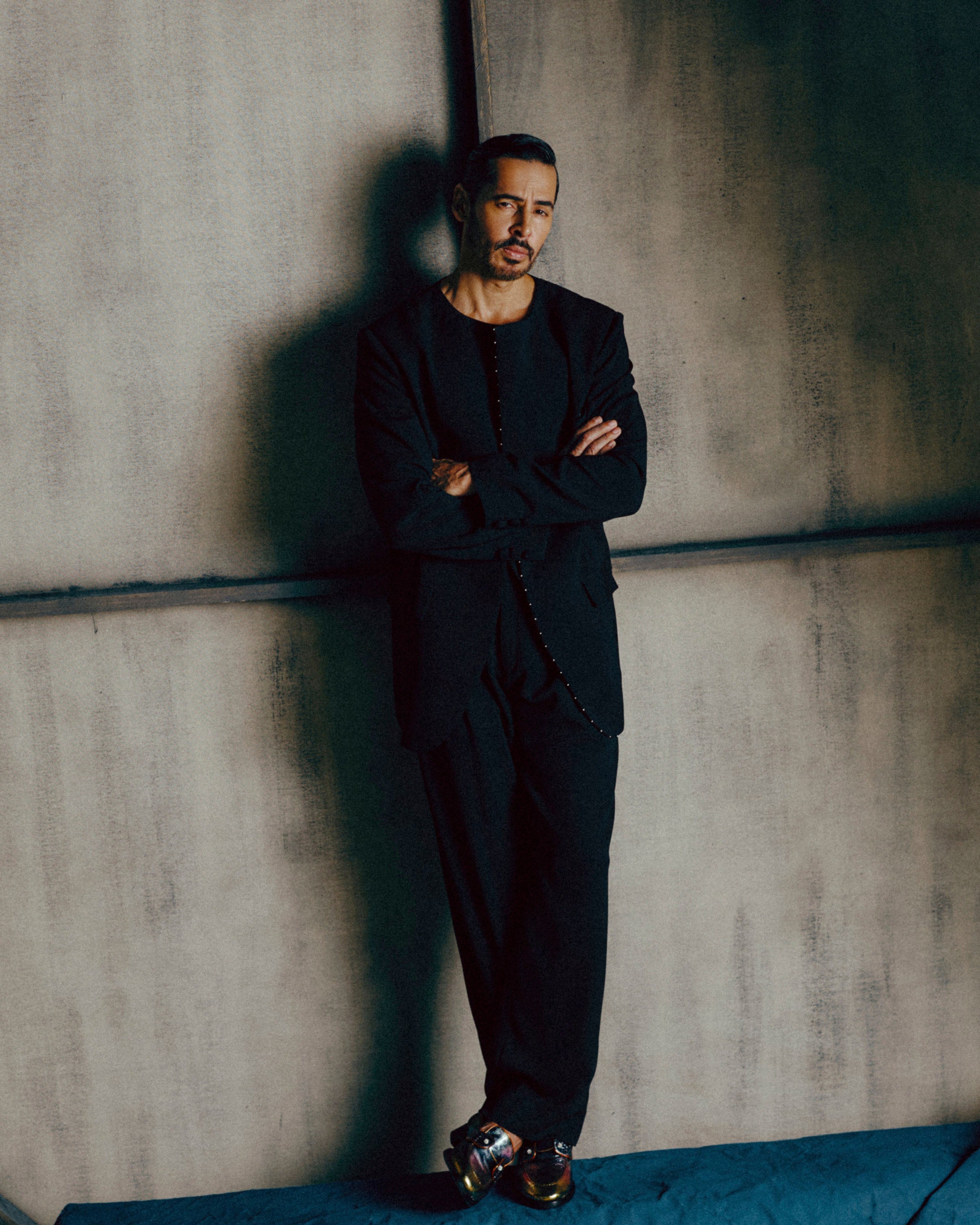 Man in a black suit standing against a textured wall with 'BAZAAR' logo.